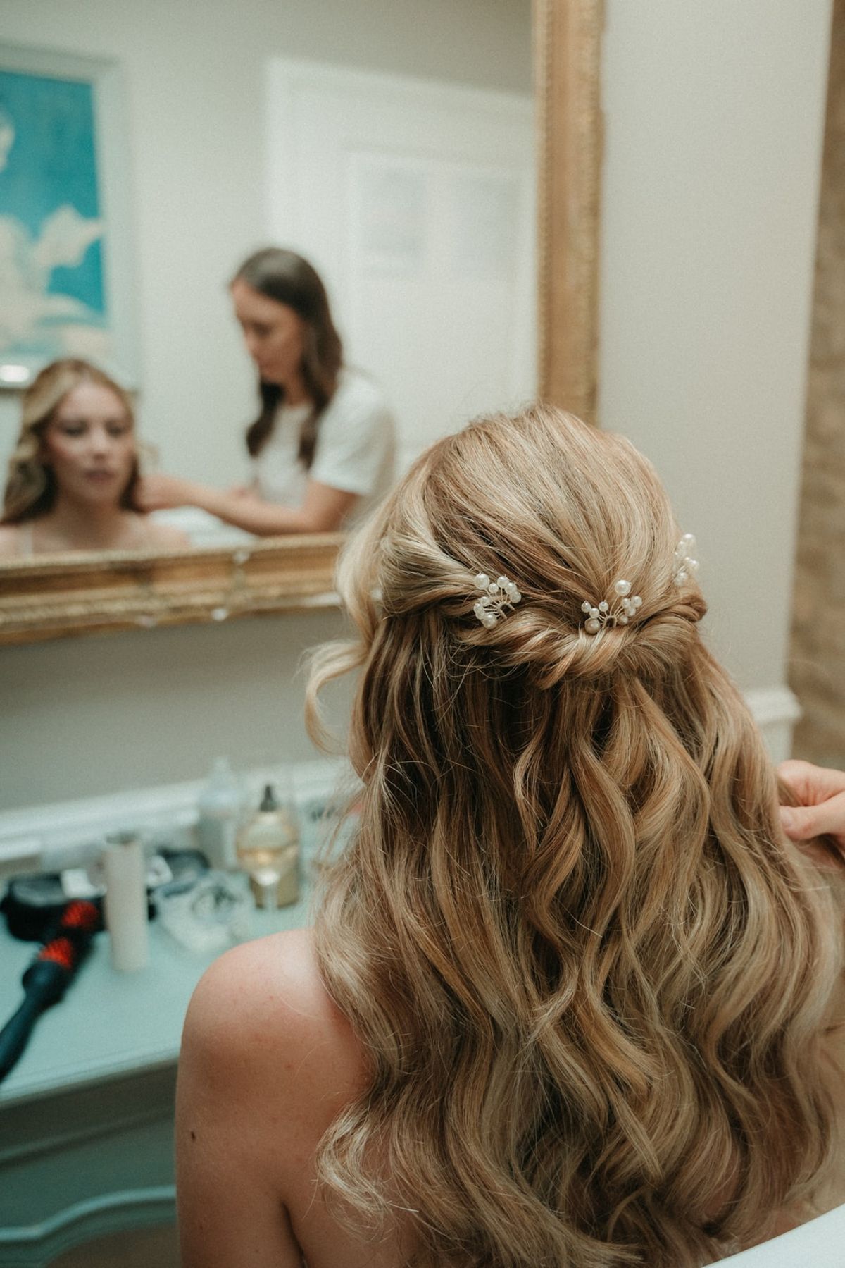 Bride from behind showing soft wavy hair with pearl floral pins, mirror reflection in vanity.