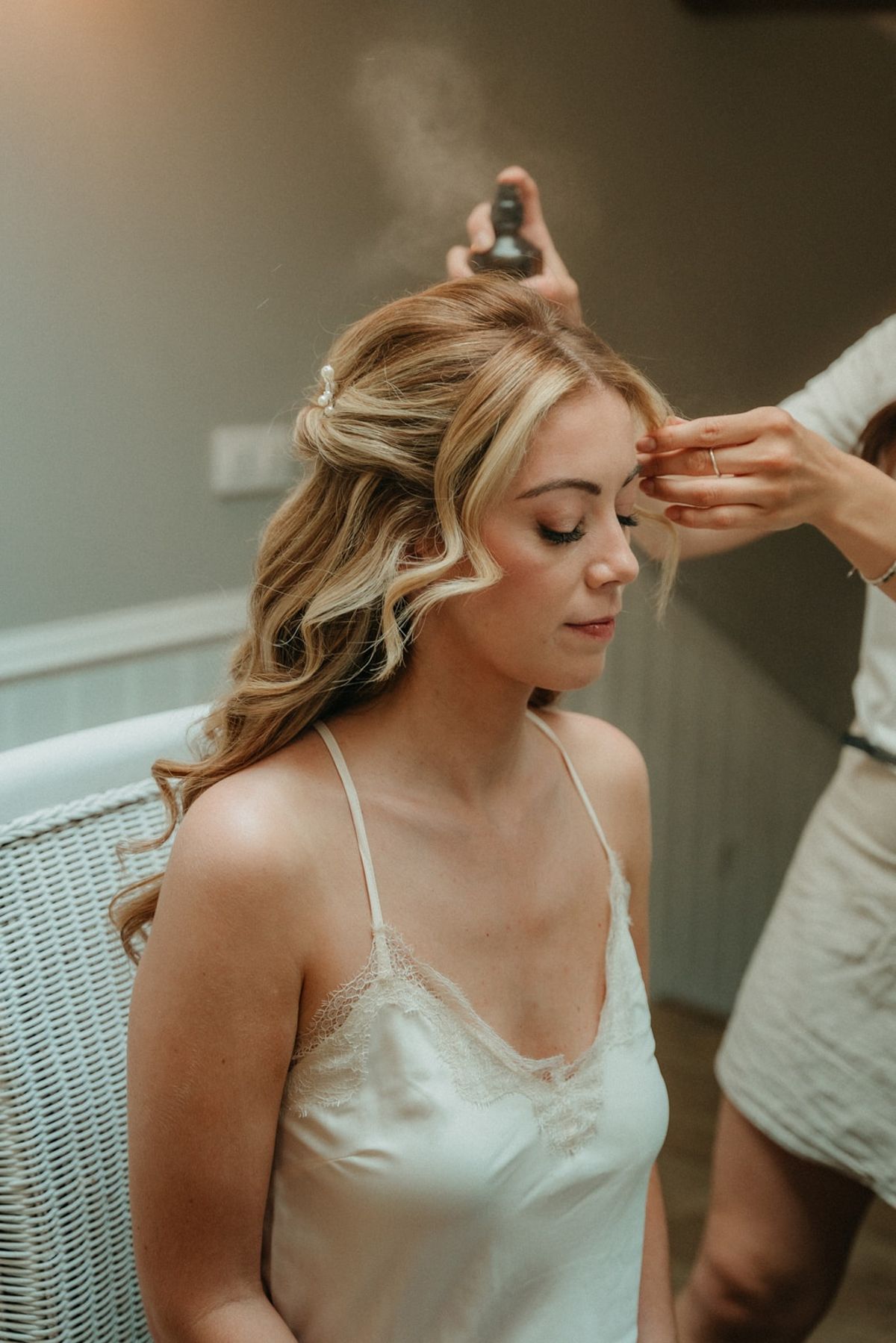 Bride in silk camisole gets hairspray applied while seated in white wicker chair.