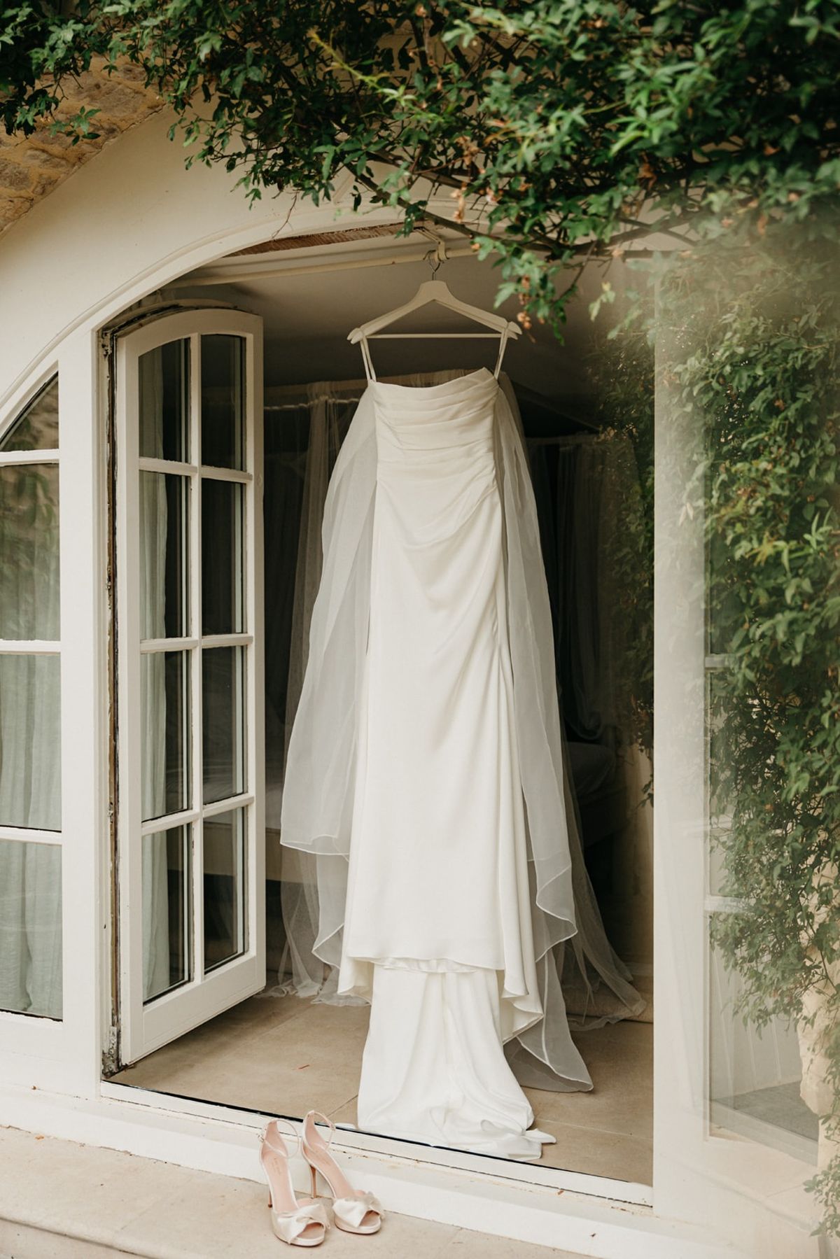 Wedding dress with tulle overlay hanging from a French door, blush heels on the stone threshold.