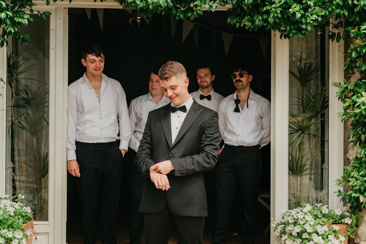 Groom adjusts cuff with four groomsmen behind in a stone doorway draped with greenery and bunting.