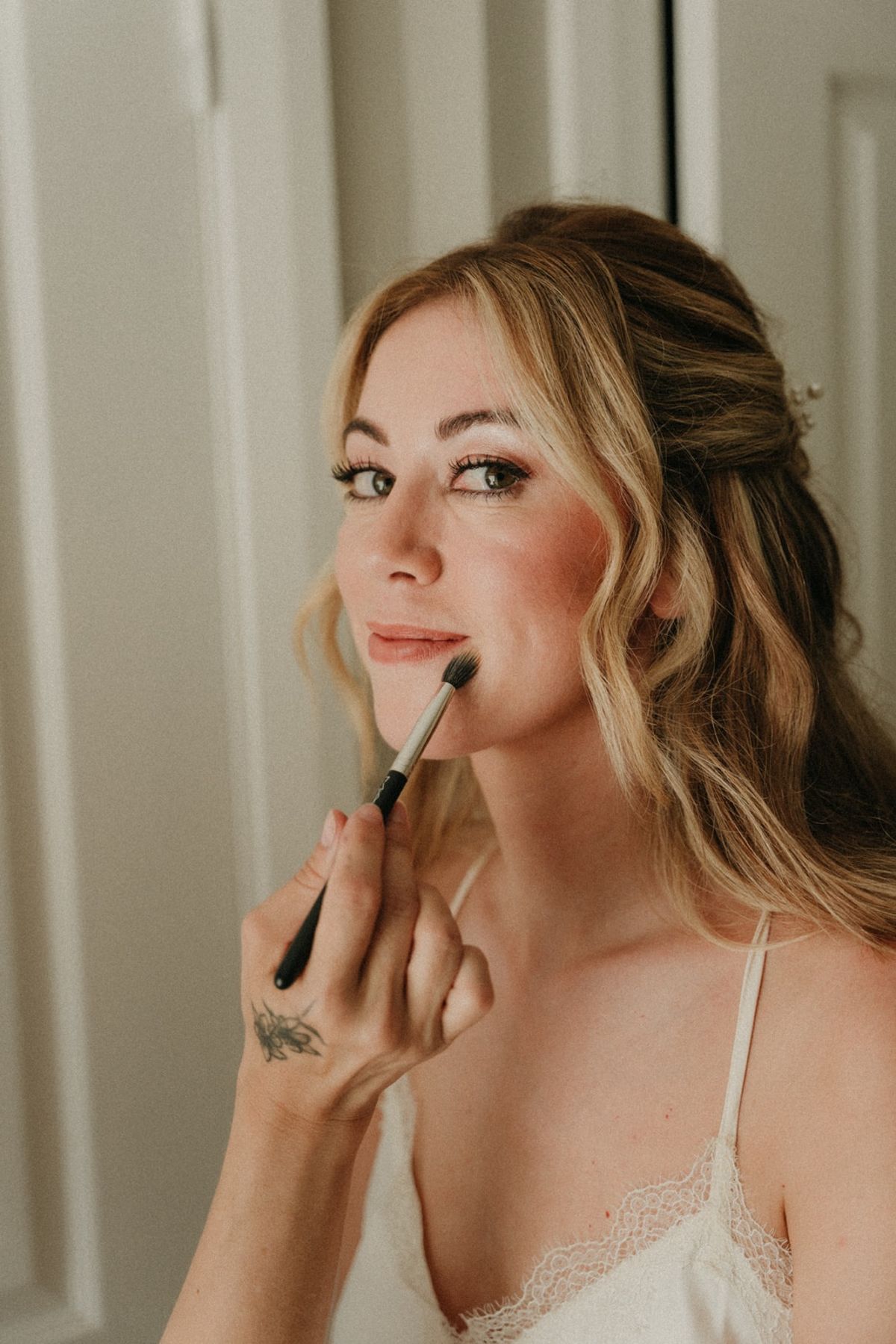 Bride mid-makeup application with brush at lip, soft waves and pearl hair clip in window light.