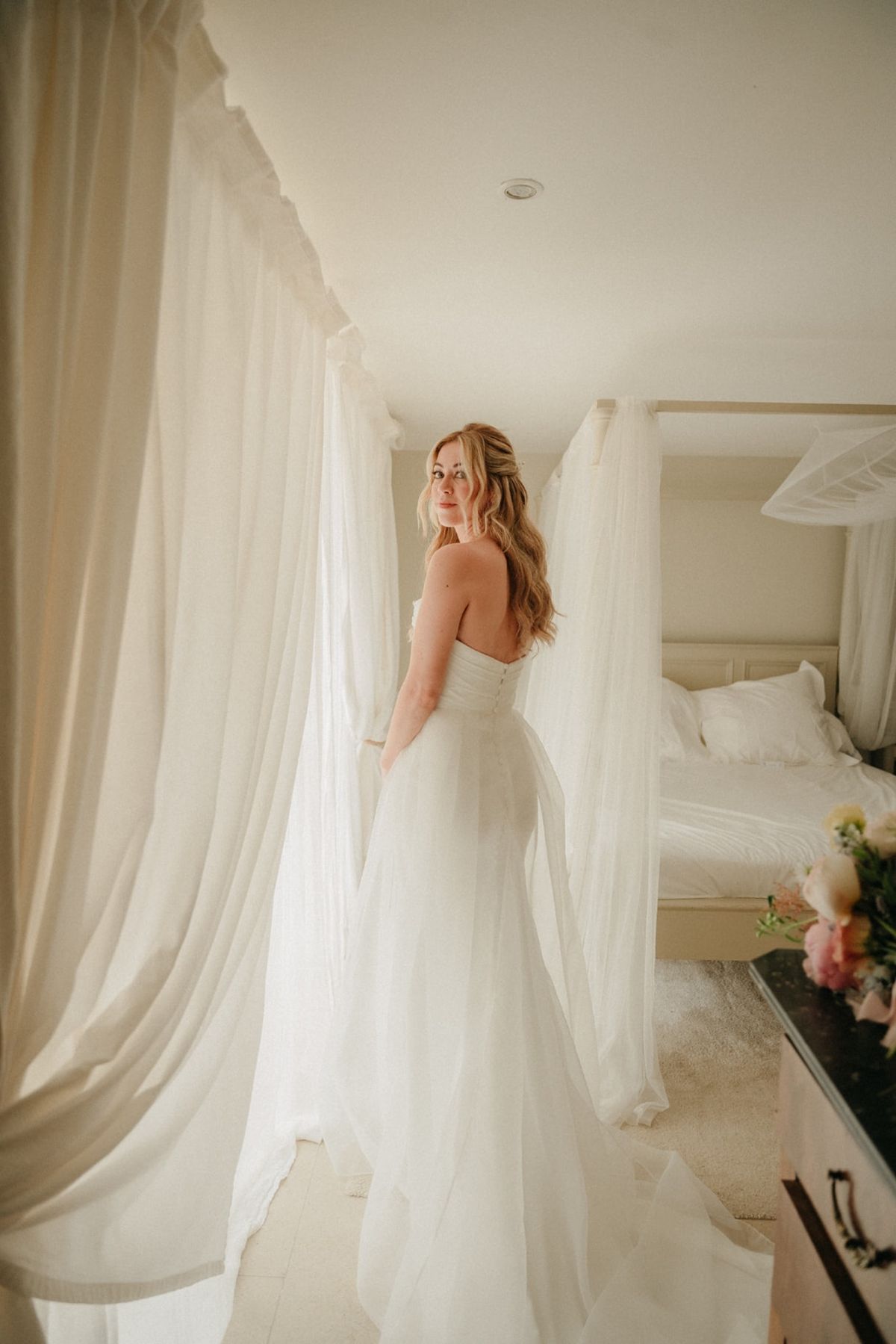 Bride in gown with tulle overlay billowing in a sunlit bedroom with sheer curtains and canopy bed.