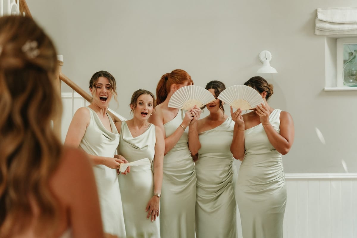 Five sage-green bridesmaids react with joy and paper fans to the bride at the first reveal.