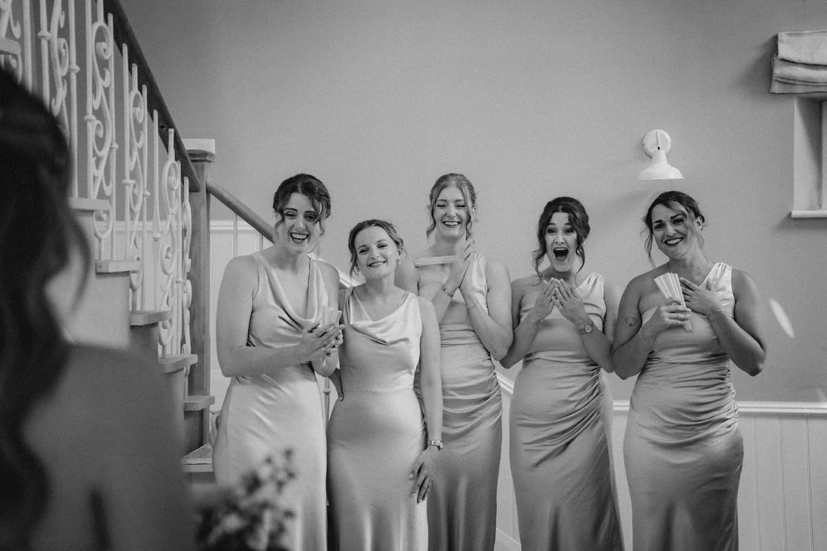 Black and white bridesmaids in sage satin laugh with raised hands seeing the bride.