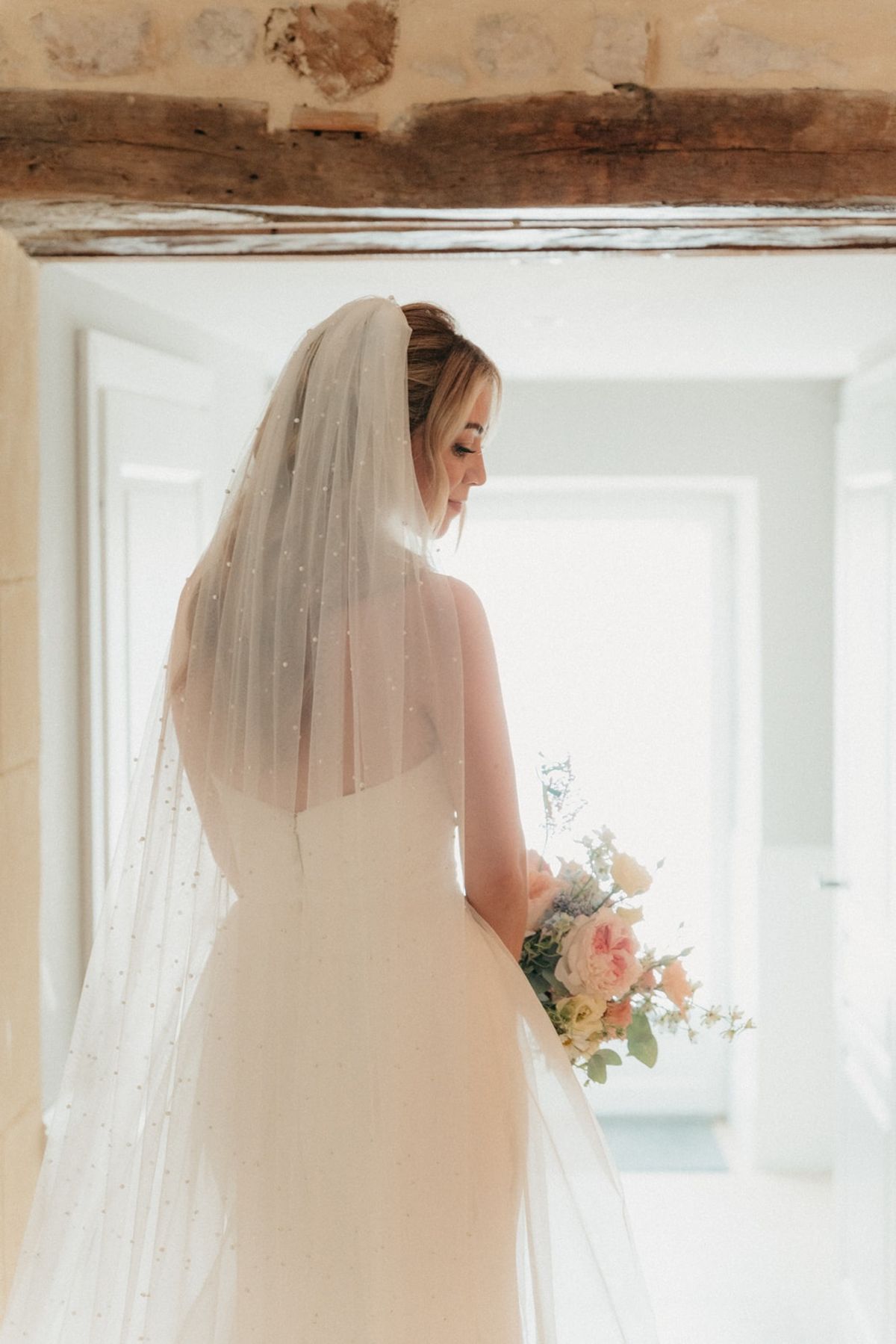 Bride looks down at pastel bouquet in stone doorway with pearl-dotted veil over shoulders.