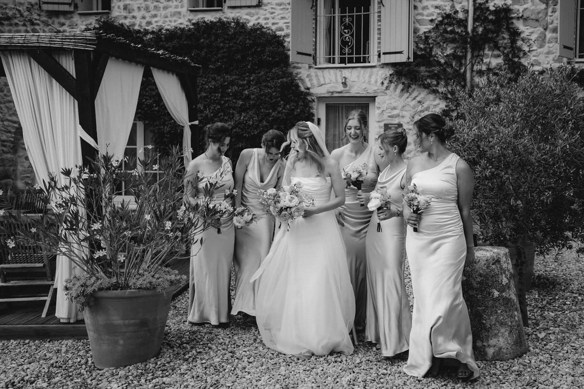 Black and white bride and five bridesmaids in sage satin walk across gravel with bouquets.