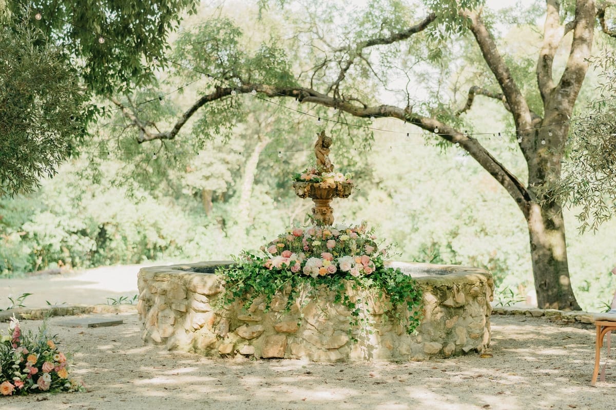 Stone cherub fountain altar dressed with cascading pastel rose florals under oak canopy.