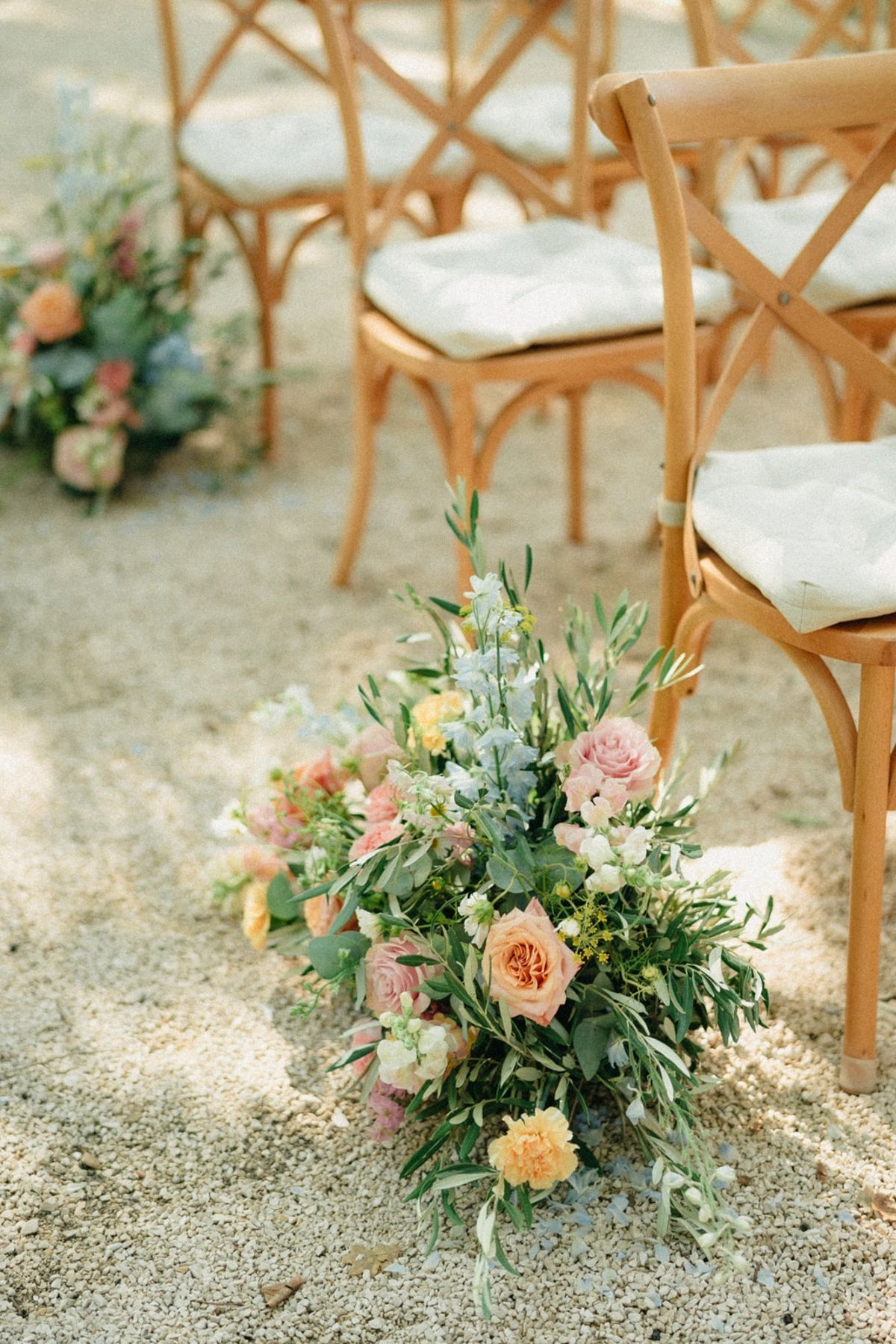 Pastel meadow-style aisle floral arrangement at the base of a wooden crossback chair.