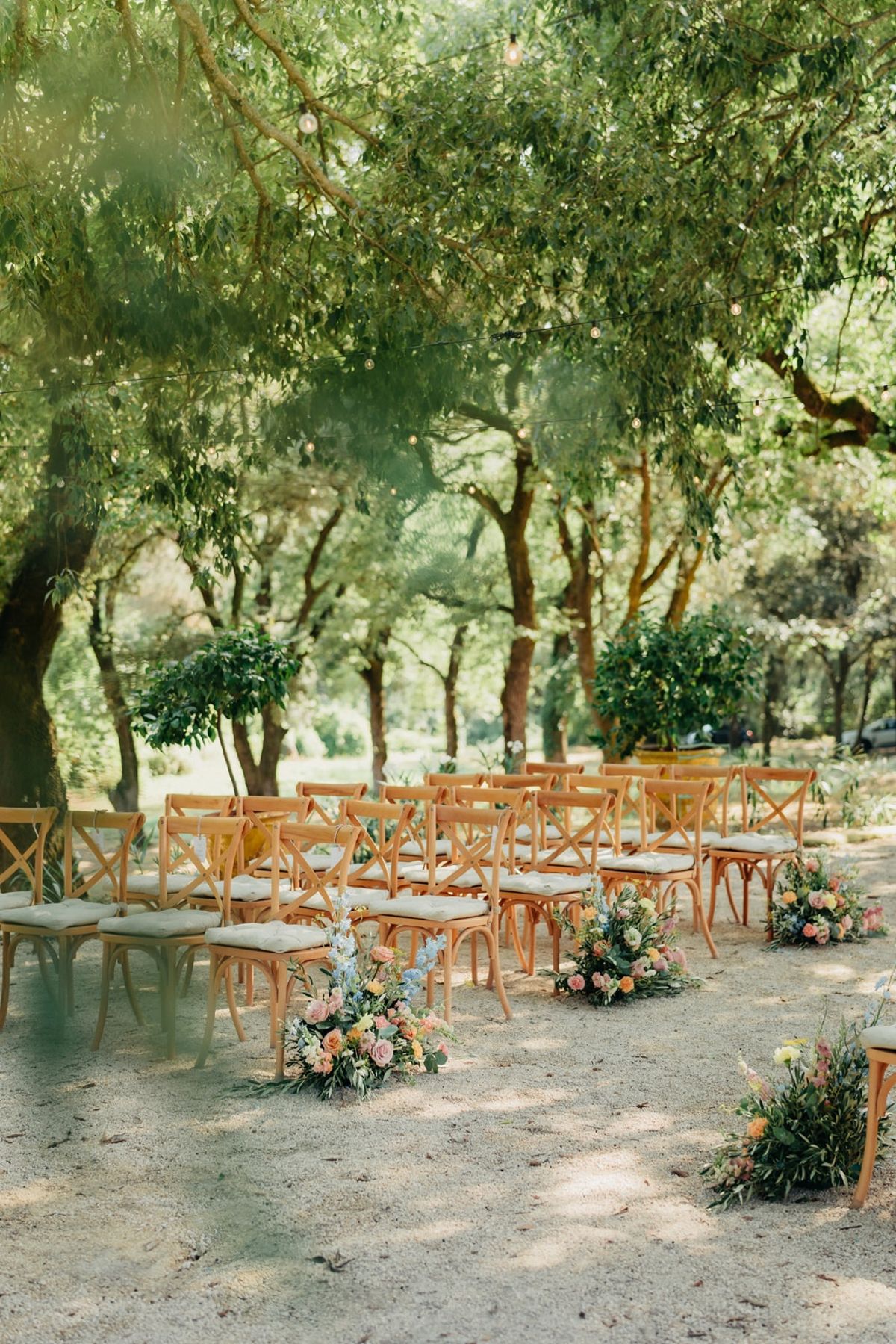 Empty crossback ceremony seating under oak canopy with meadow florals down the gravel aisle.