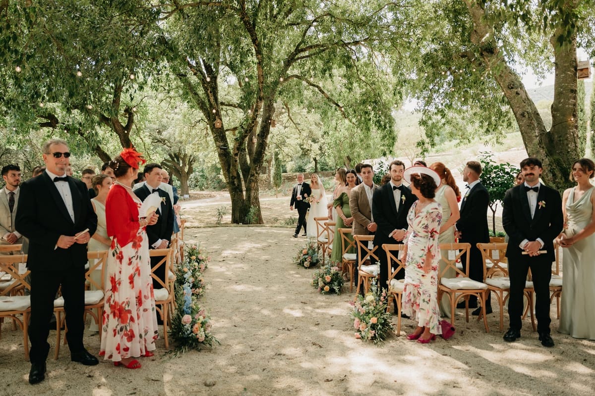 Guests stand at outdoor ceremony as couple gathers at stone fountain altar with mountains beyond.