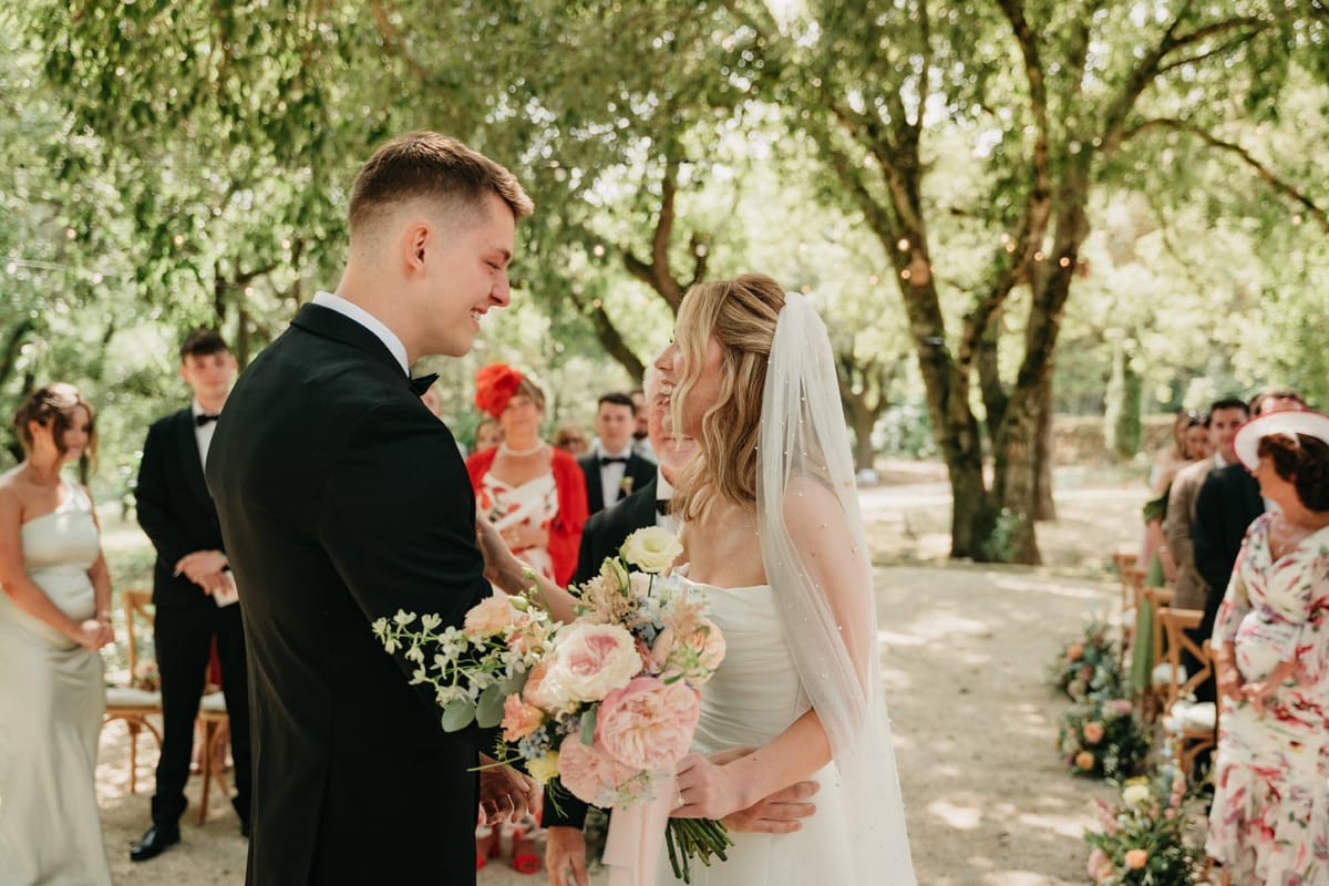 Bride and groom smile facing each other at the ceremony start with pastel bouquet and oak canopy.