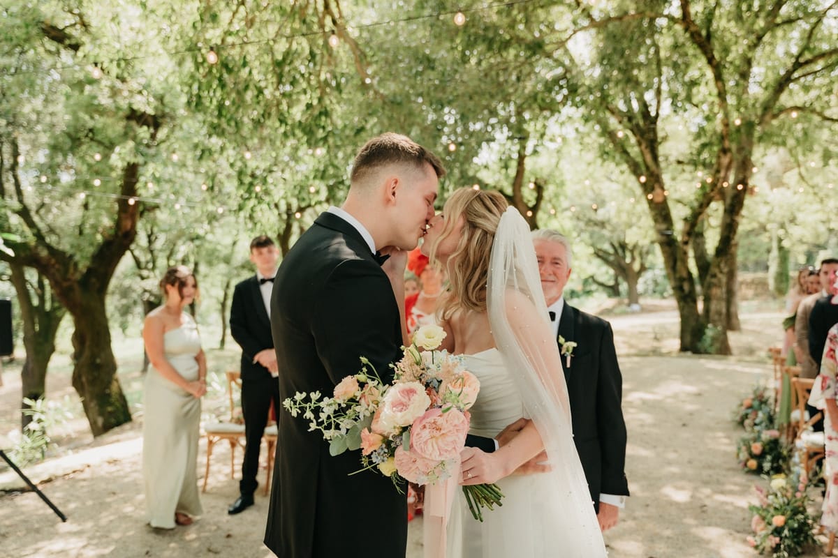 Newlyweds share first kiss under oak canopy with officiant and guests on either side.