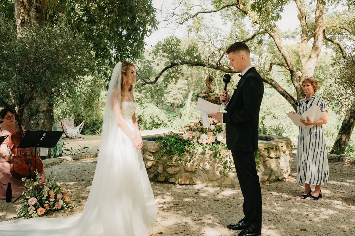 Groom reads vows from card with bride listening, officiant and string quartet through the trees.