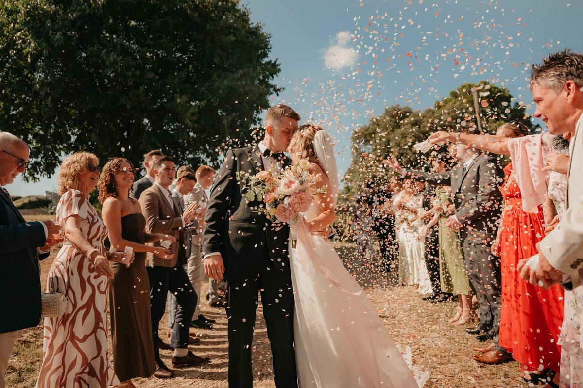 Couple kiss inside swirl of confetti petals at the ceremony exit with cheering guests.