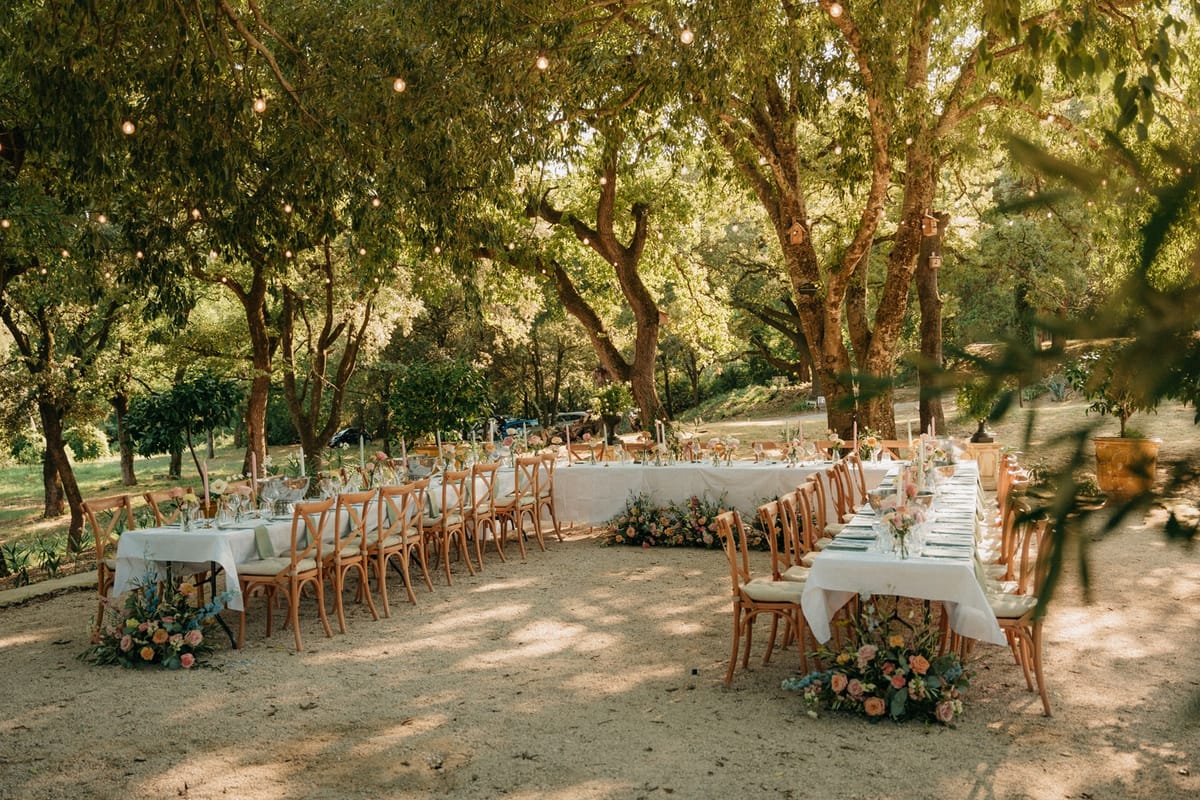 Empty open-air horseshoe banquet under oak trees with white linens, candles and pastel florals.