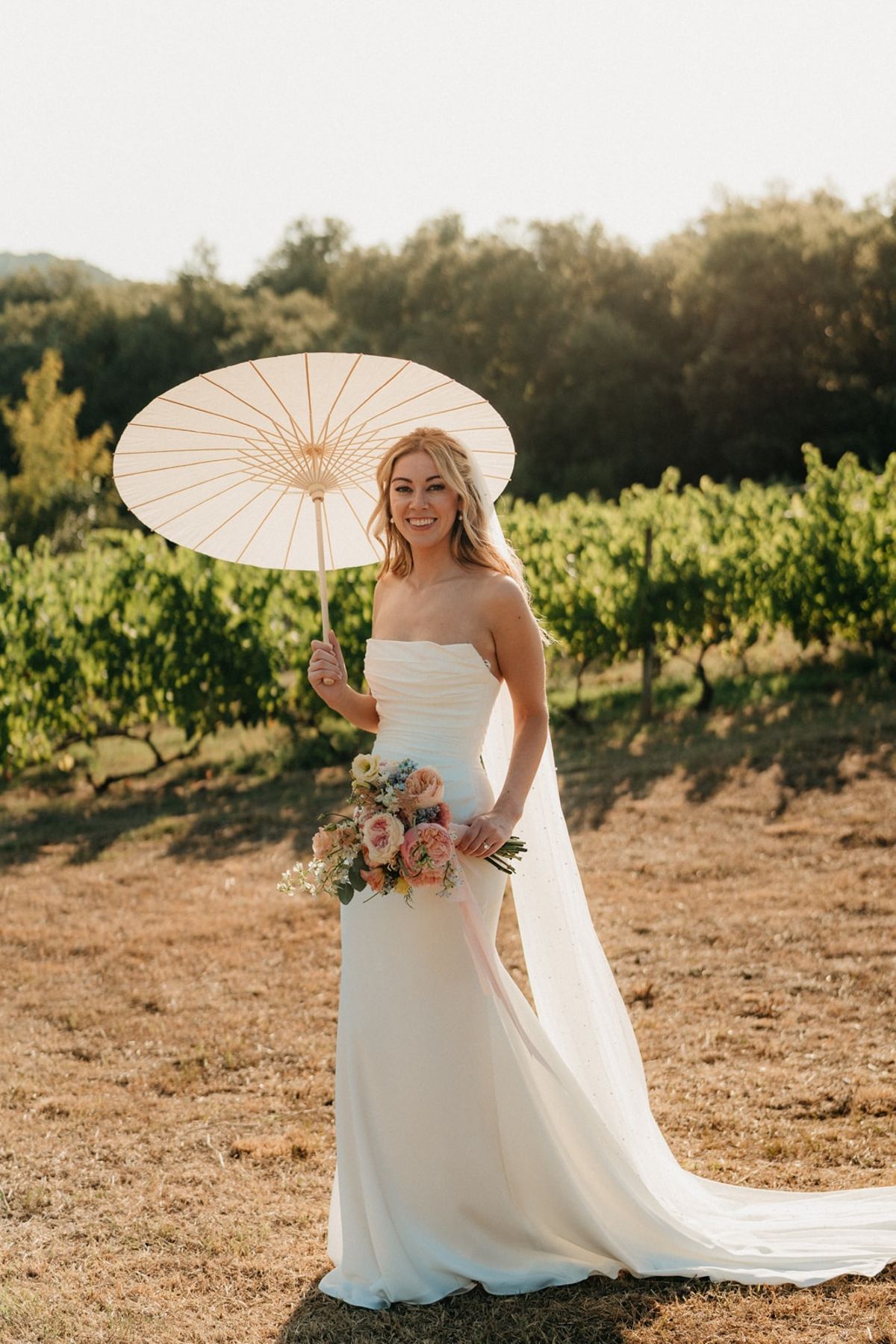Bride in vineyard with paper parasol and pastel bouquet in warm golden hour light.
