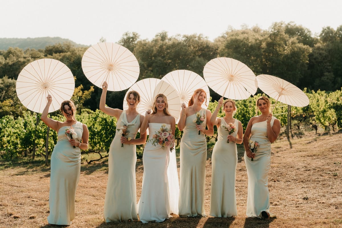 Bride and five sage-clad bridesmaids hold paper parasols and posies in a sunlit vineyard.