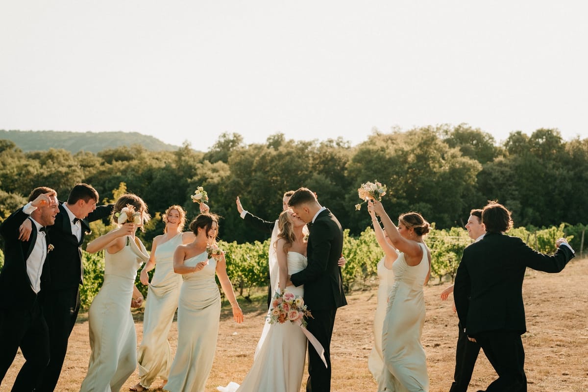 Couple kiss centred in a vineyard huddle of cheering bridesmaids and groomsmen.