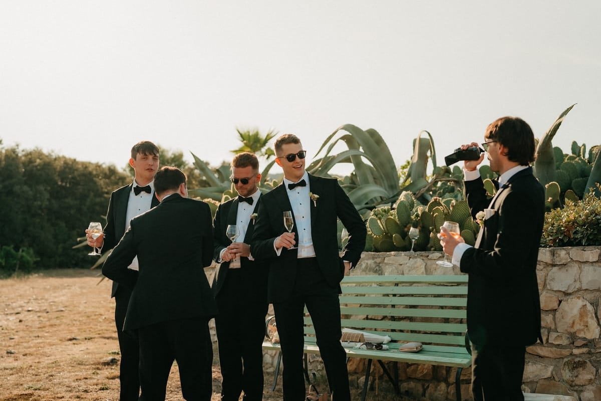 Groomsmen with champagne on cactus terrace in golden light, one in dark sunglasses.