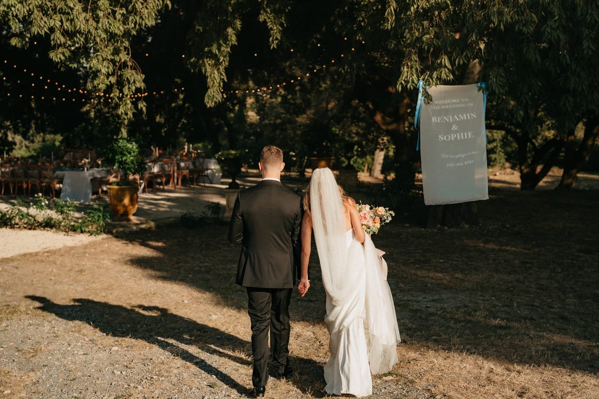 Back view of couple approaching painted welcome sign with festoon-lit dining beyond.