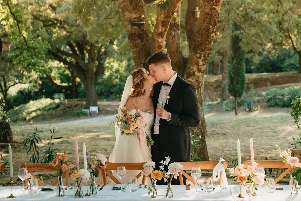 Couple kiss at head of long dining table with pastel florals and taper candles under oak trees.