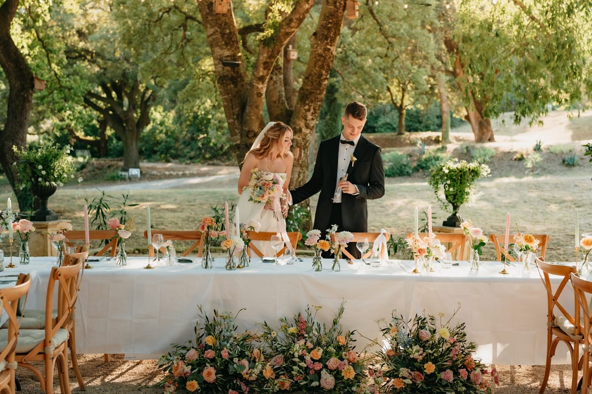 Couple at long table head with raised flute, lined with bud florals and meadow border at base.