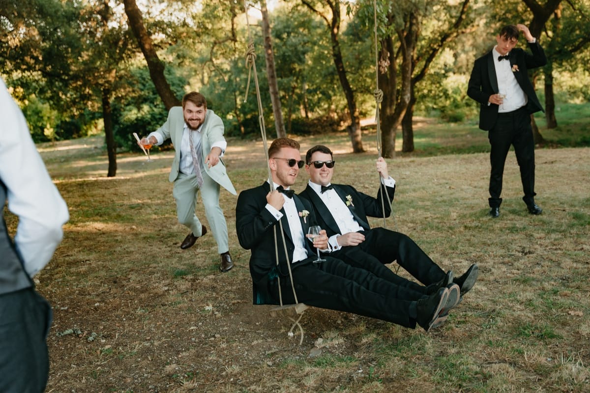 Groomsmen push two friends on a tree swing with an aperitif mid-air during cocktails.
