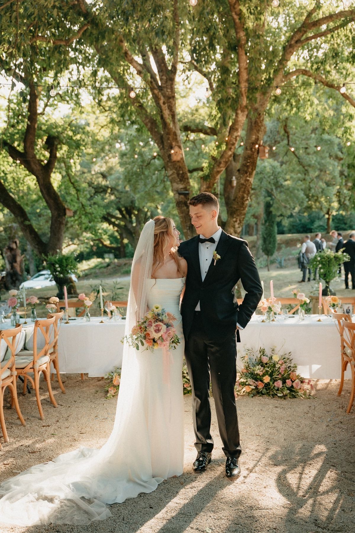 Couple stand at dining clearing entrance with festoon lights and guests beyond in golden light.