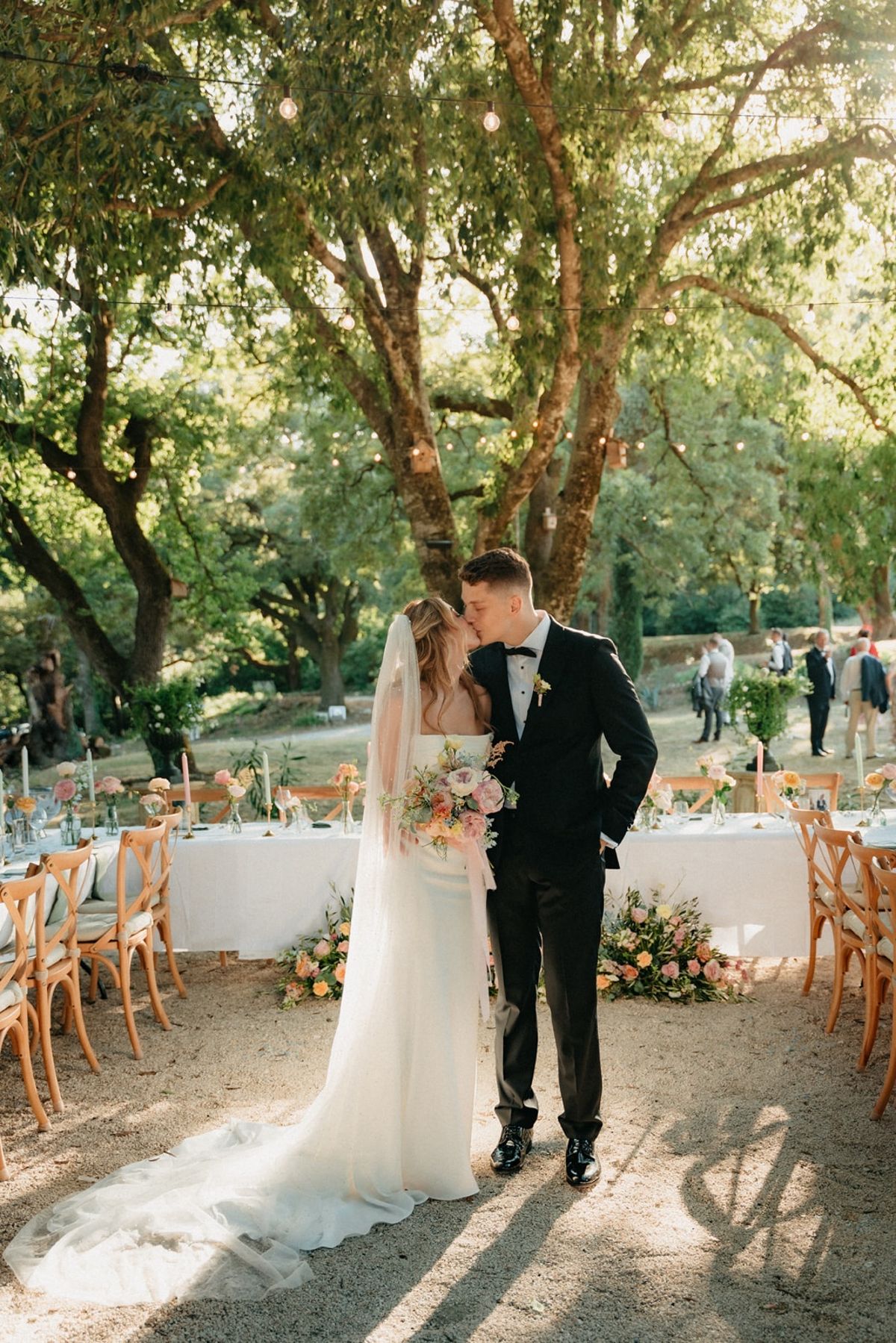 Couple kiss at entrance to open-air banquet with festoon-lit oaks and dressed tables behind.