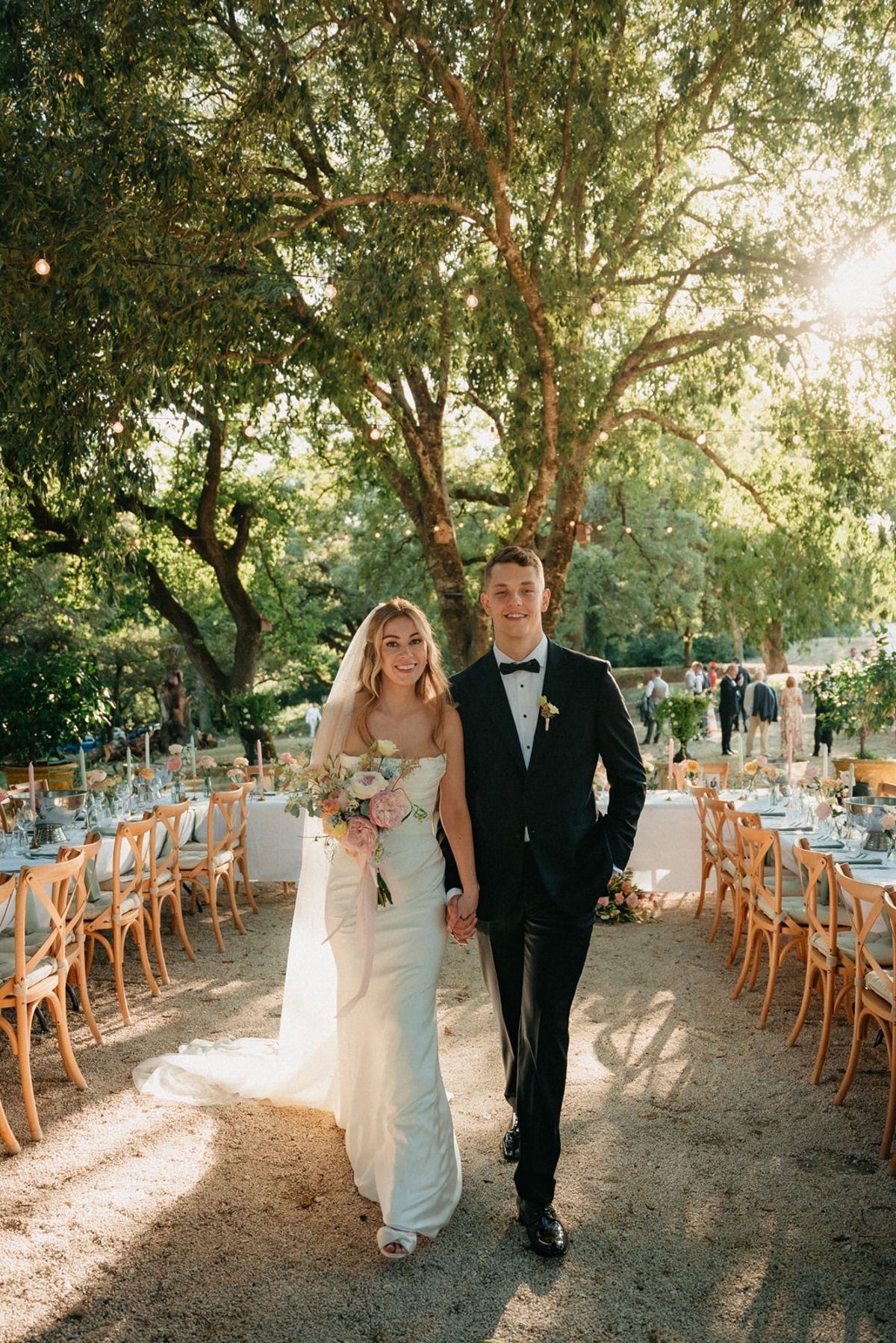 Couple walk hand in hand between long dressed tables in the open-air banquet aisle.