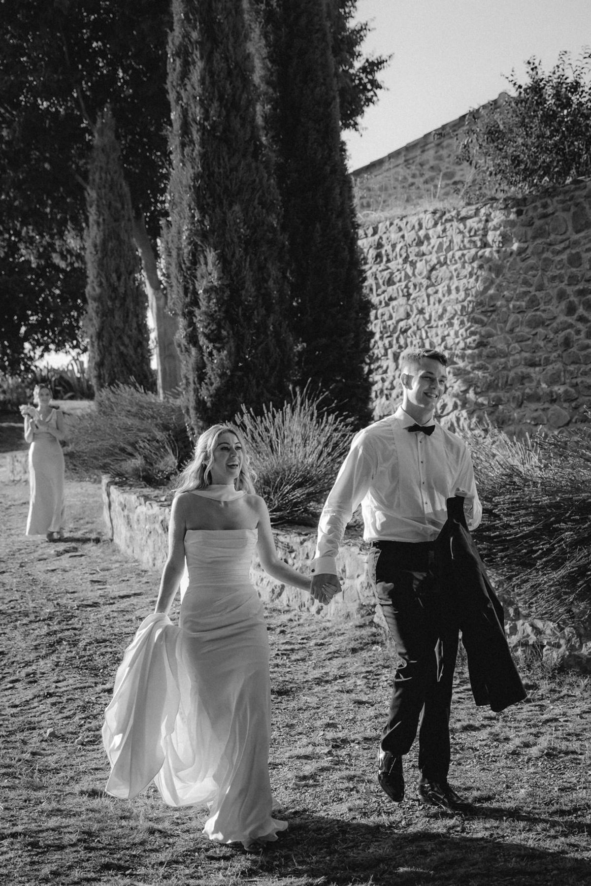 Black and white couple walk hand in hand past stone wall and cypress, jacket over groom's arm.