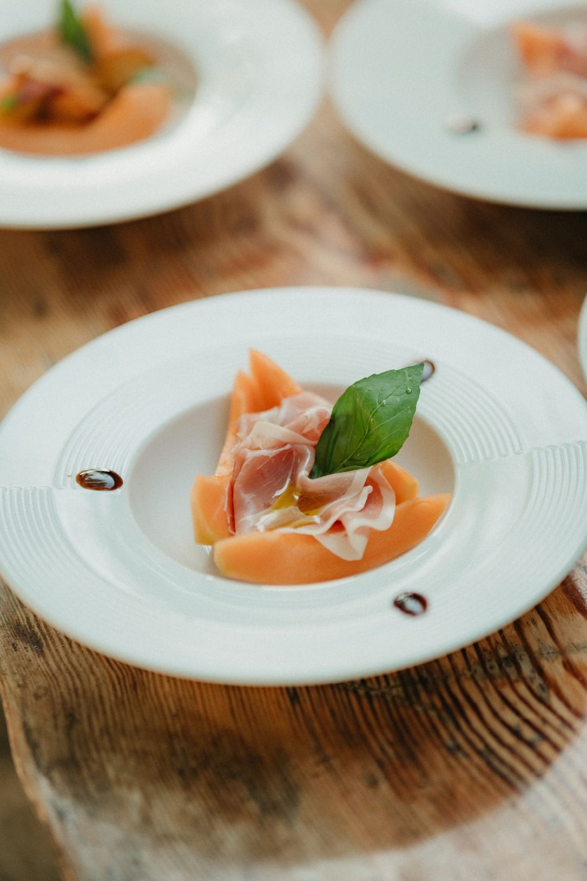 Plated melon and prosciutto first course with basil and balsamic on rustic wooden table.