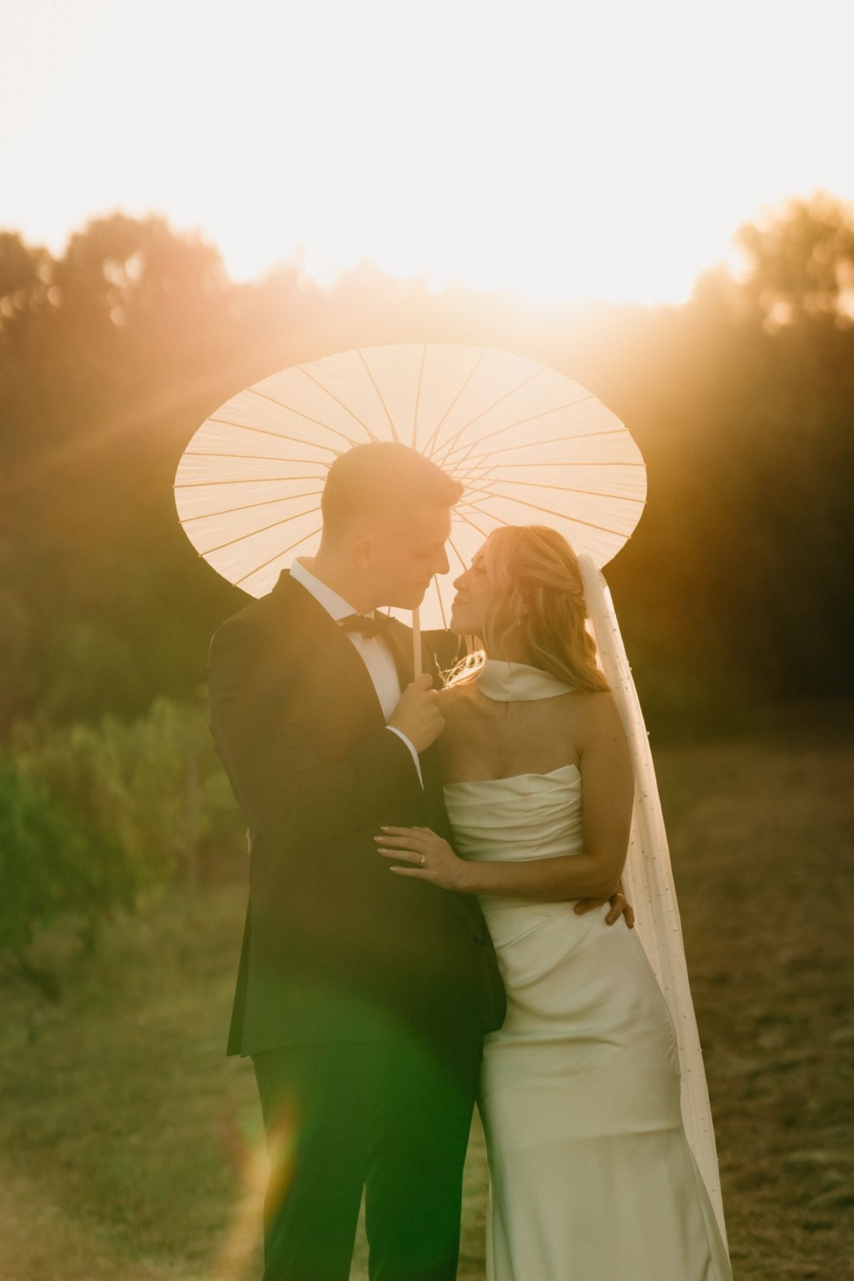 Backlit golden hour couple portrait under paper parasol in a vineyard with sun rays around.