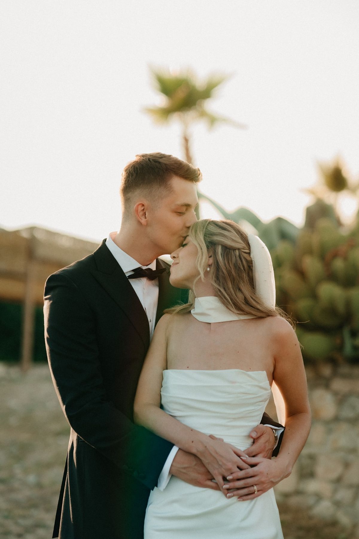 Groom kisses bride's forehead with white neck scarf, palm and cactus garden softly behind.