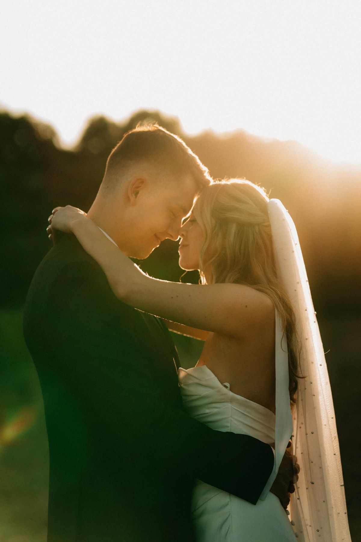 Backlit couple embrace forehead to forehead in meadow with veil catching golden sunset.