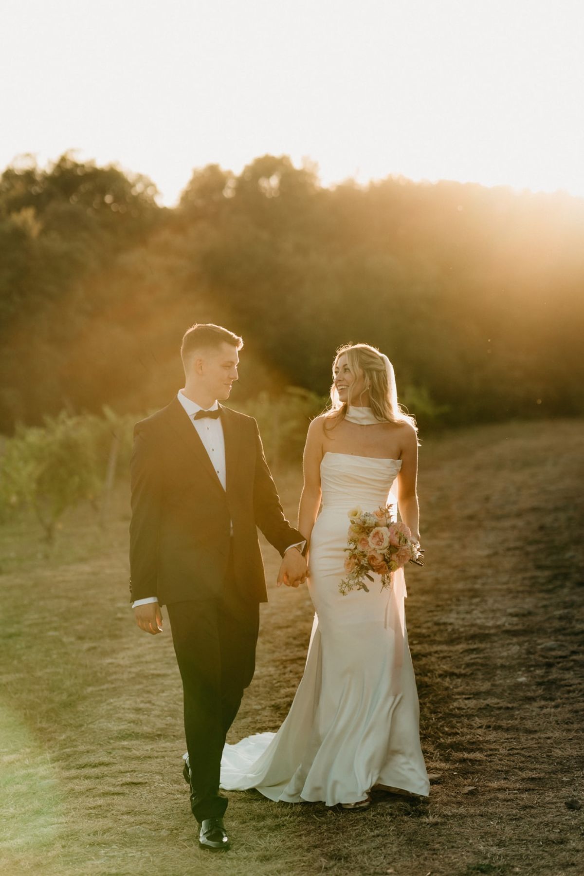 Couple walk hand in hand through sunlit meadow with pastel bouquet and low sun flare behind.