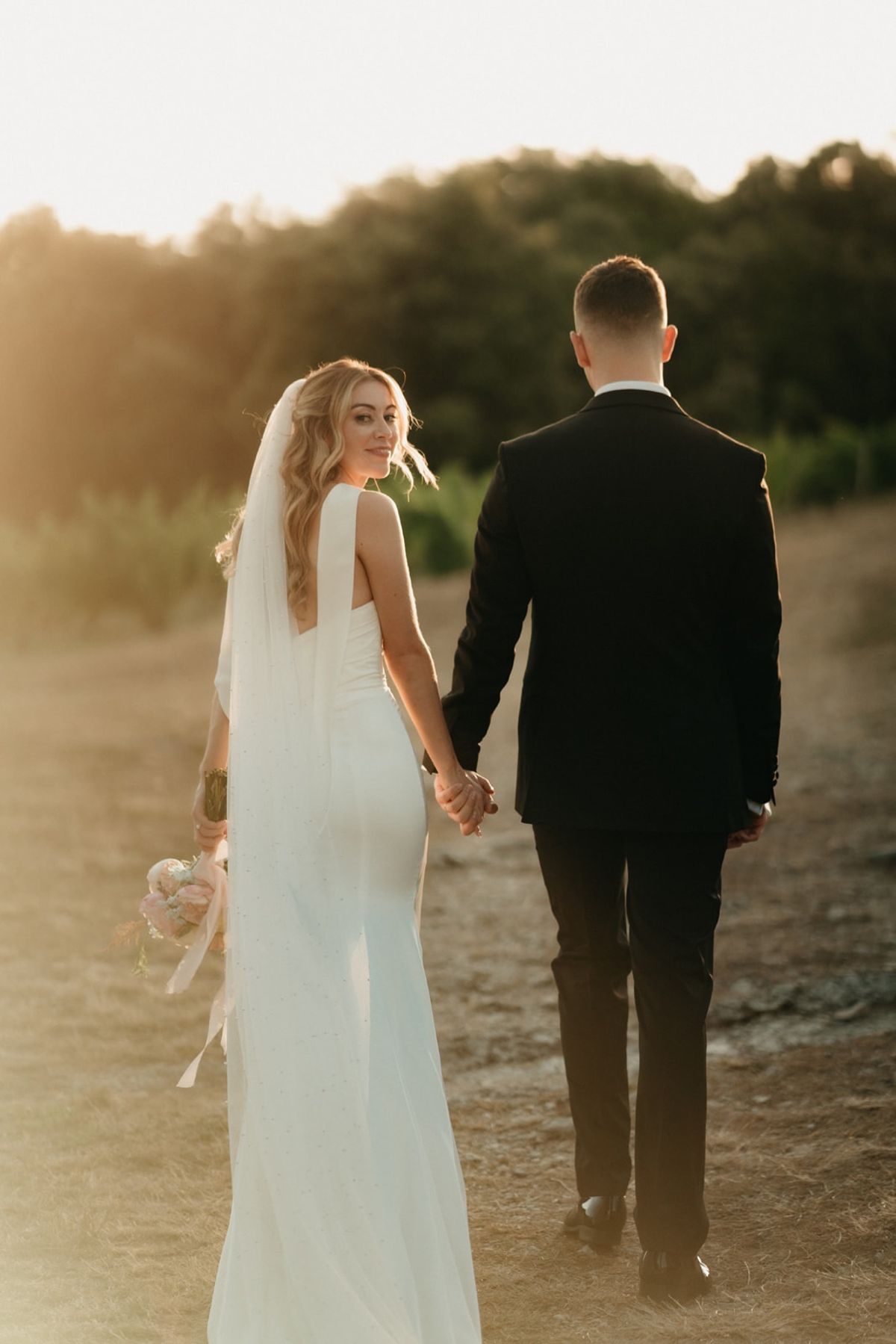 Couple walk away on sunlit vineyard path, bride glances back over shoulder with bouquet.