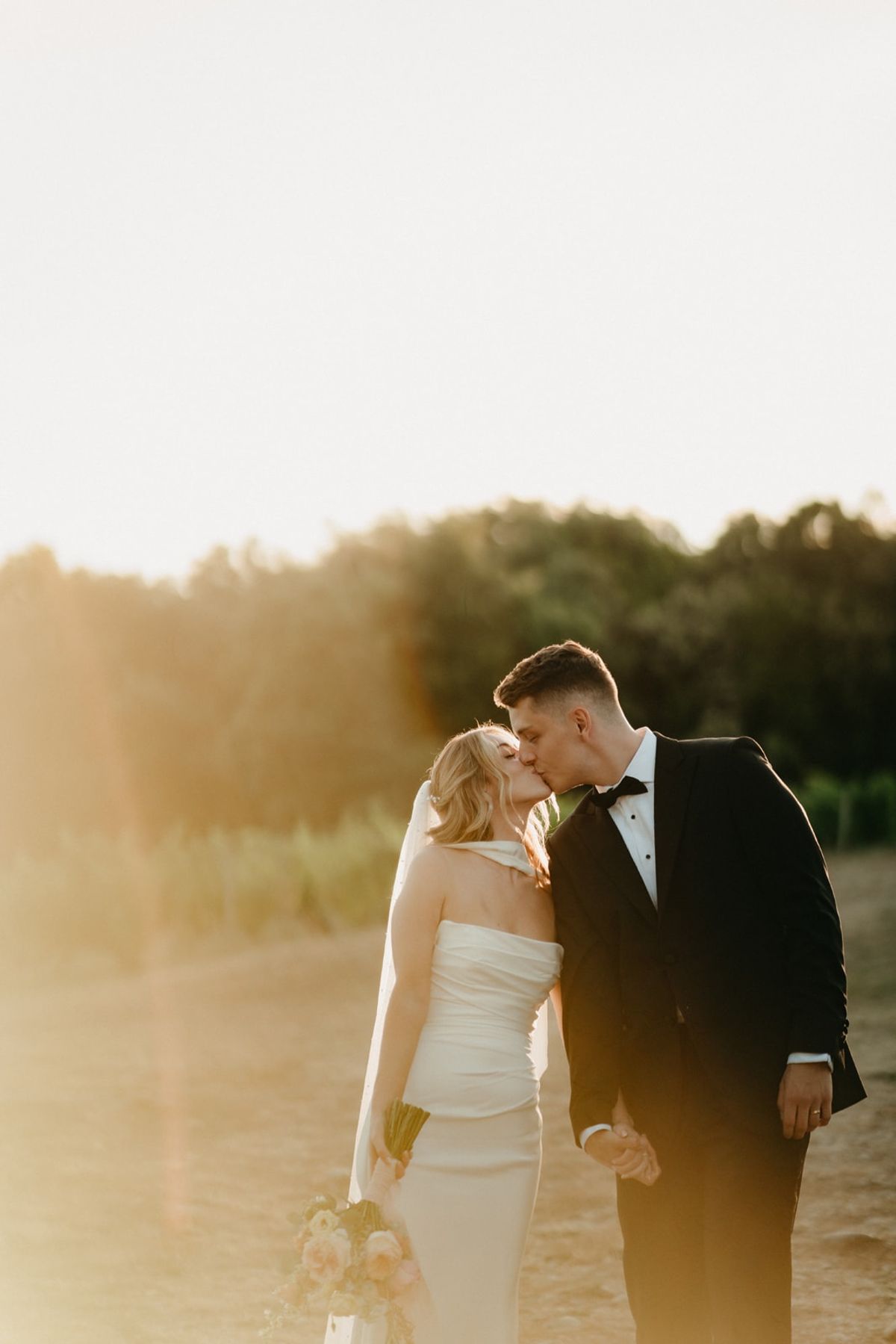 Backlit couple kiss in sunset flare with bride's white neck scarf catching the warm light.