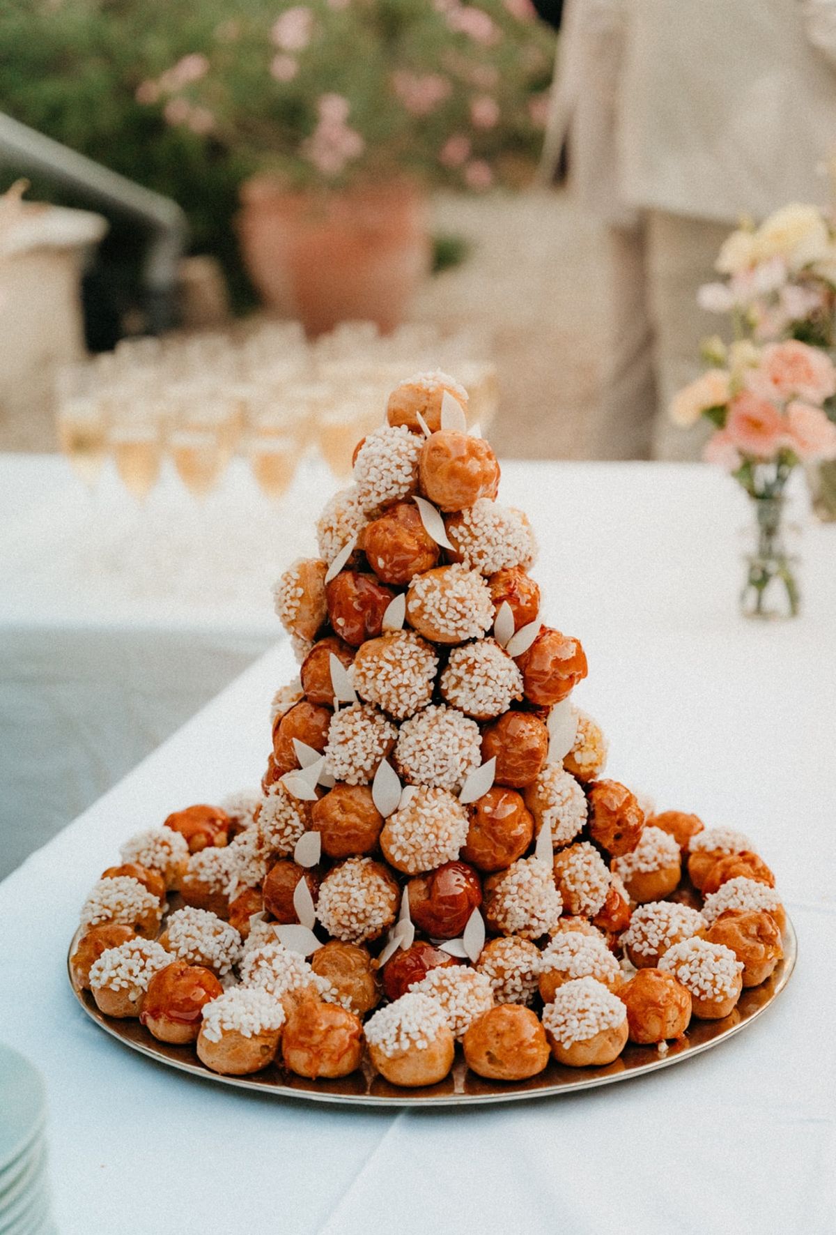 Croquembouche tower with crystallised sugar pearls and rows of champagne flutes behind on linen.