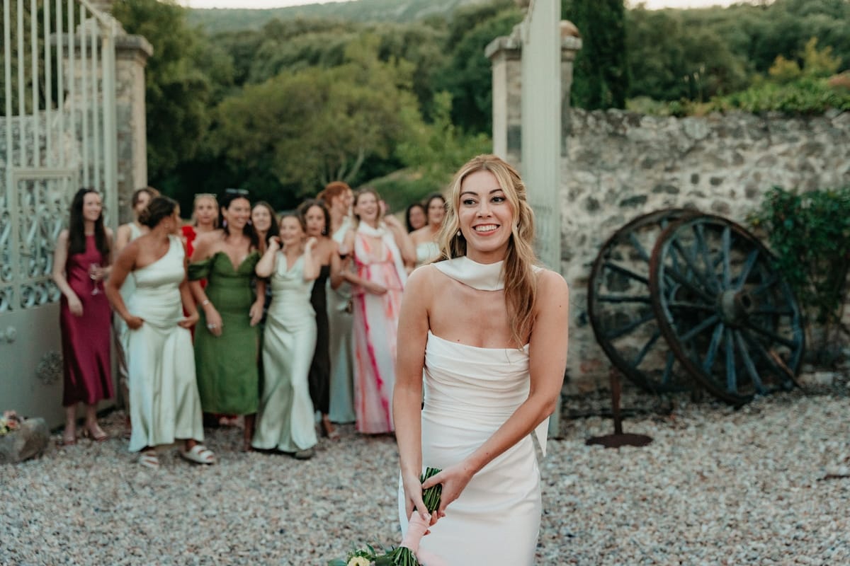 Bride at stone gates ready to throw bouquet, women guests wait behind with old cartwheel beyond.