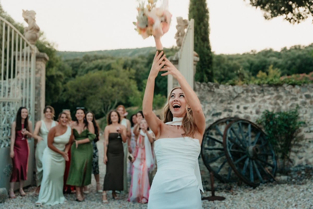 Bride throws pastel bouquet overhead with raised arms as women guests wait behind her at gates.