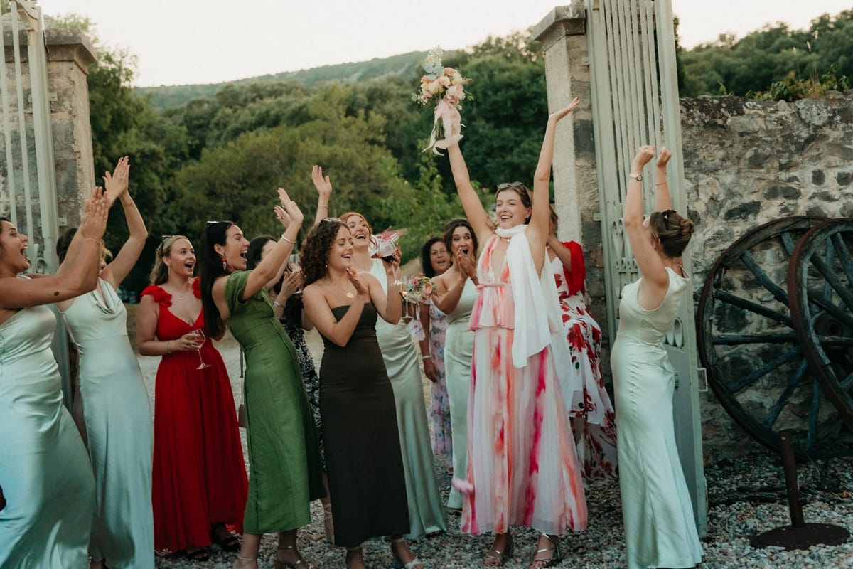 Guest in pink dress catches pastel bouquet with raised arms as women guests cheer around her.