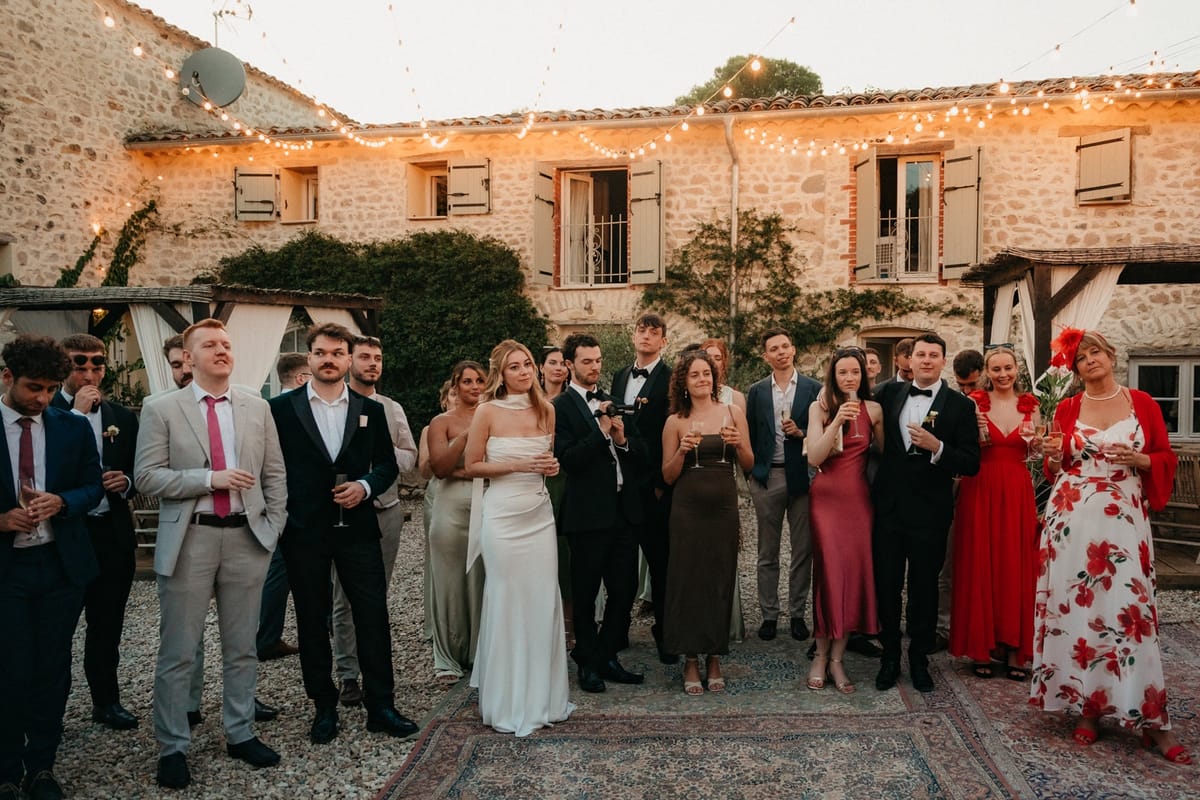 Guests gather in festoon-lit stone courtyard at dusk watching speeches with farmhouse behind.