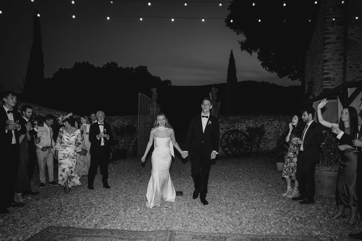 Black and white couple grand entrance into courtyard through stone gates at dusk with cheering guests.