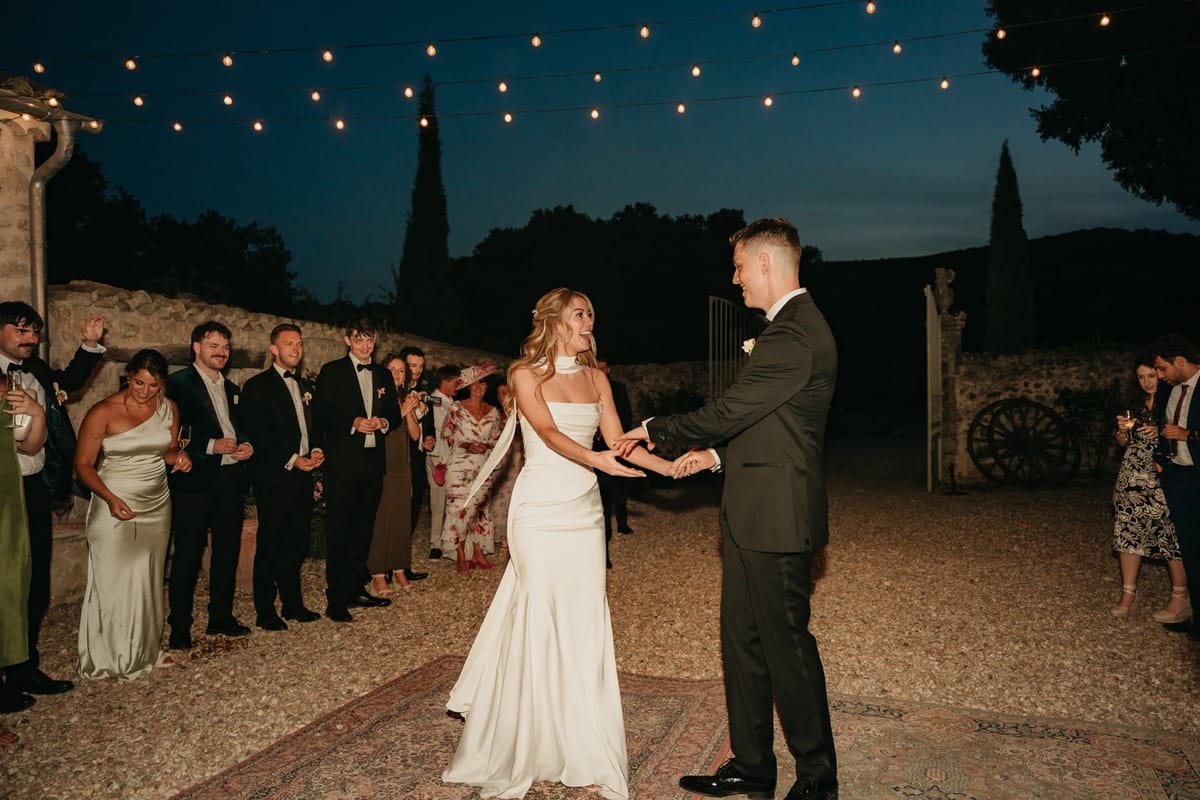 Couple's first dance in stone courtyard under festoon lights with white satin neck scarf.