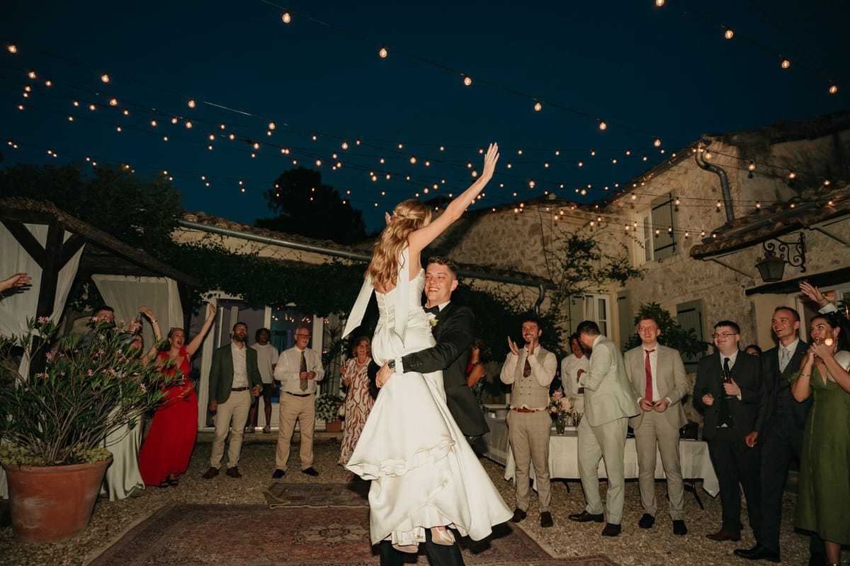 Bride lifted off the ground in joyful first dance under festoon lights with cheering guests.