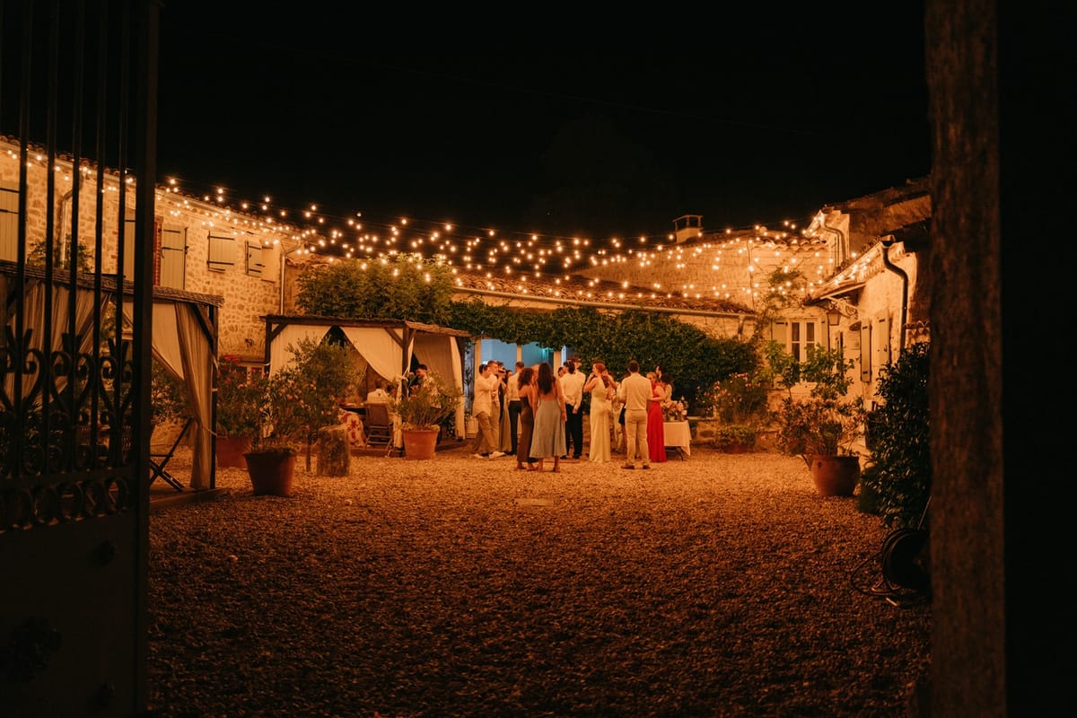 Wide night view through stone gates into courtyard with warm festoon lights and mingling guests.