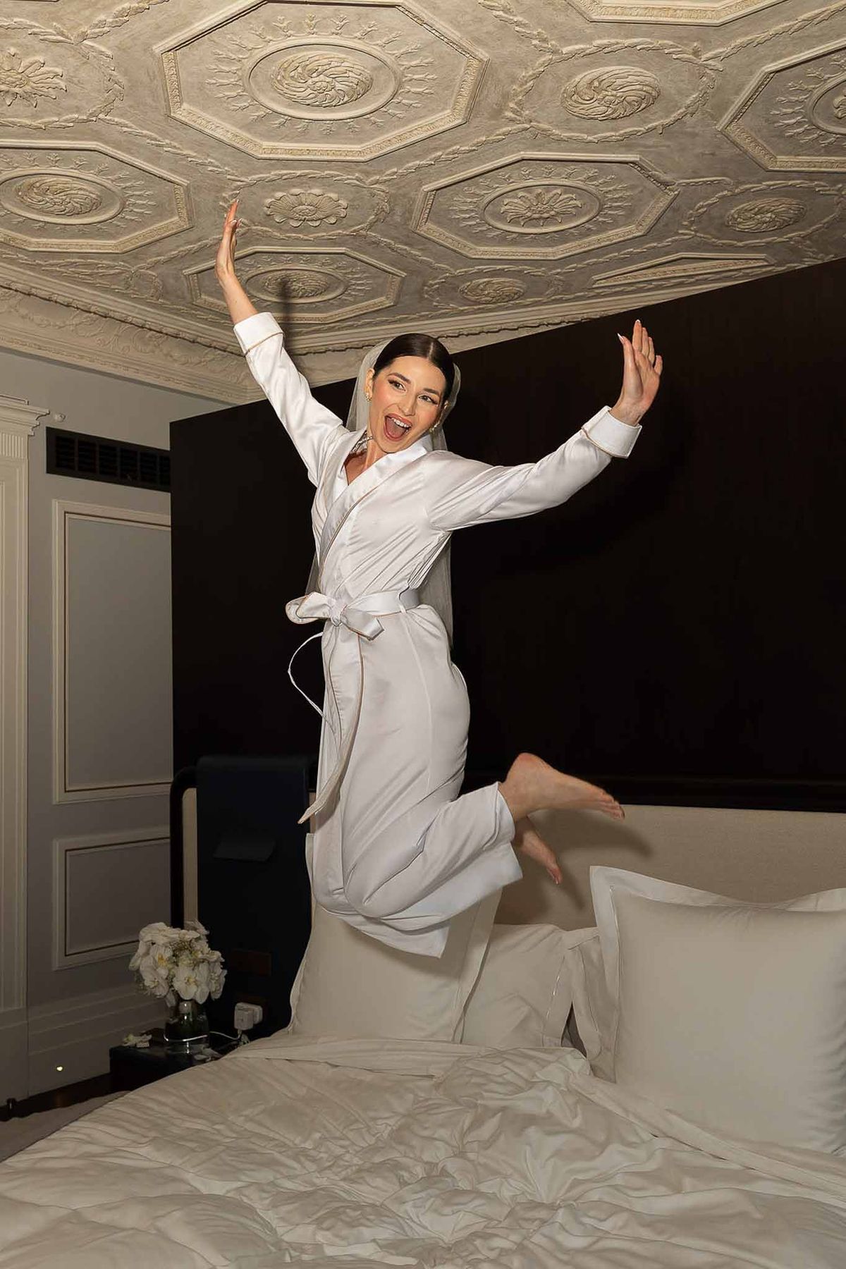 Bride in white satin robe jumping on bed in ornate chateau room with coffered ceiling during getting ready