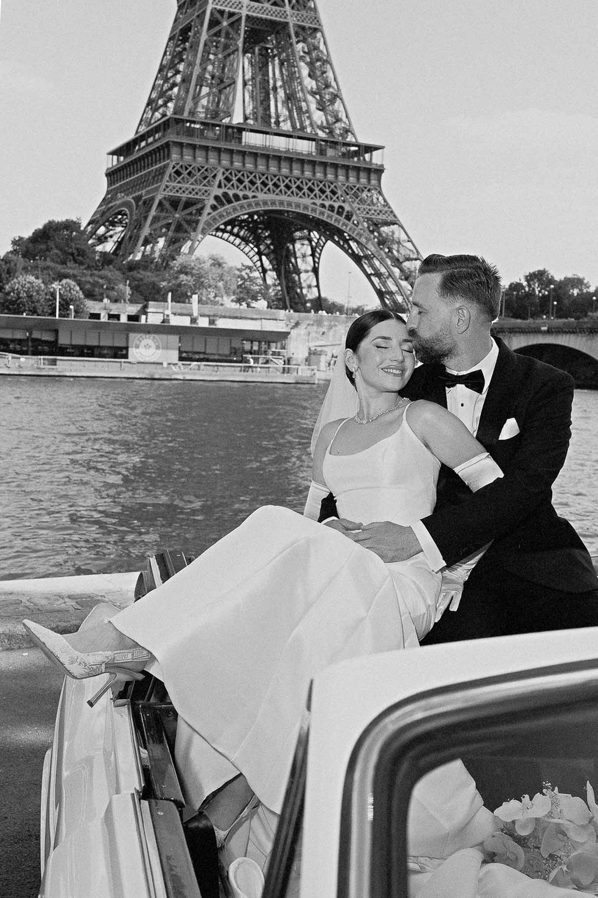 Black and white groom kissing bride on Seine with Eiffel Tower behind bride in satin gown with cathedral veil on convertible