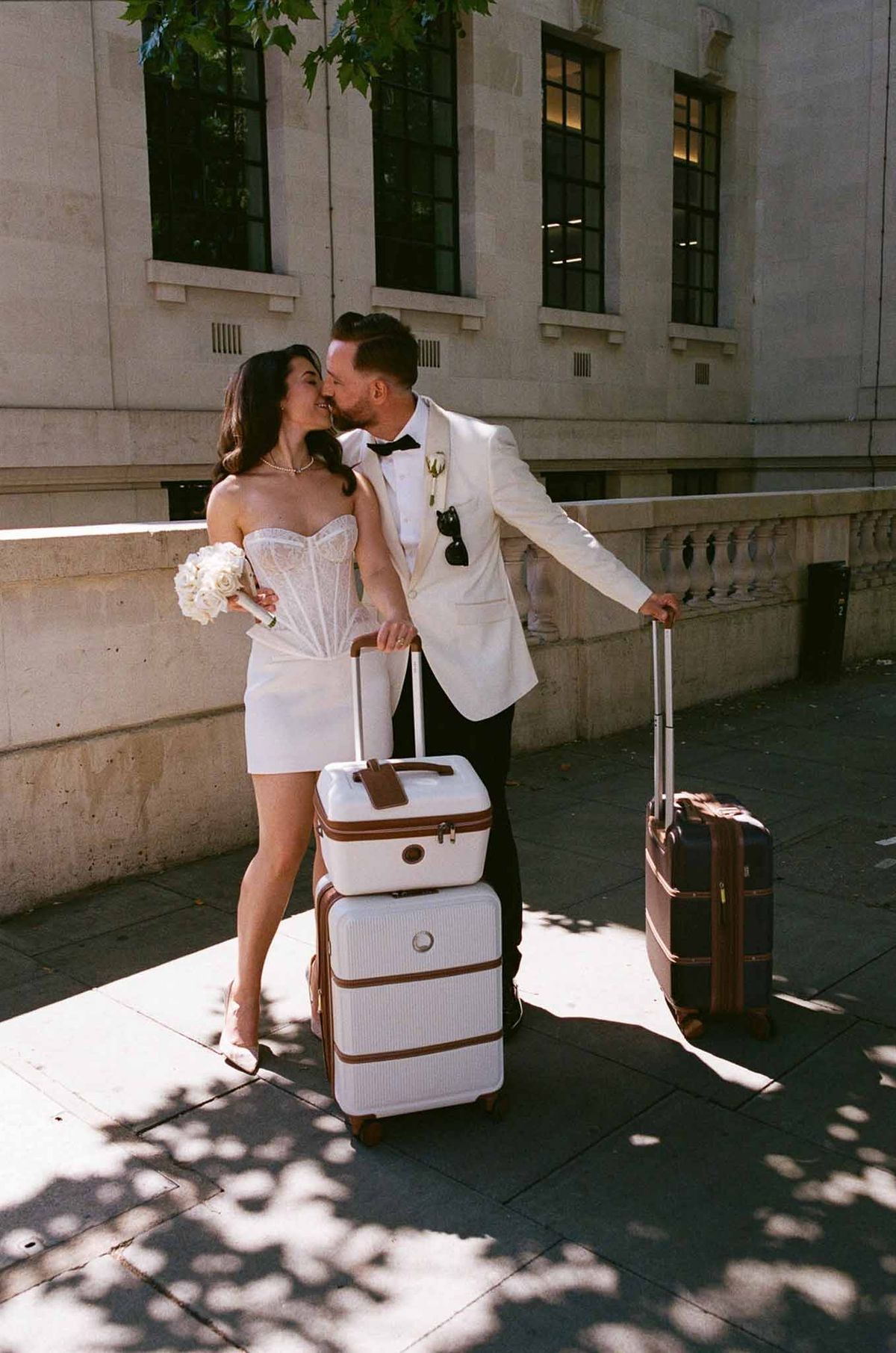 Bride and groom kissing on city pavement with vintage luggage, bride in white mini skirt and lace top